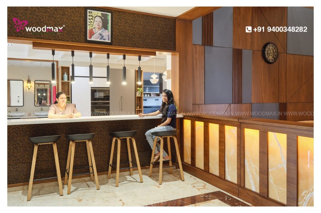 Woodmax Interiors designed the kitchen with a refrigerator where a woman is currently standing, possibly in Kasaragod, Mangalore or Kerala.
